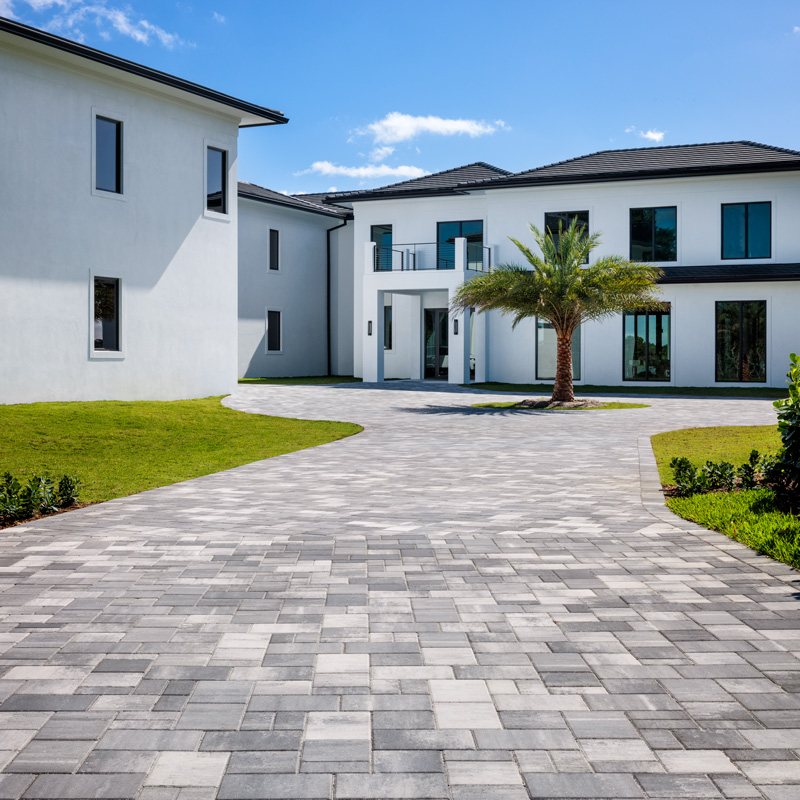 Wide gray paver driveway leading to a modern two-story home with a palm