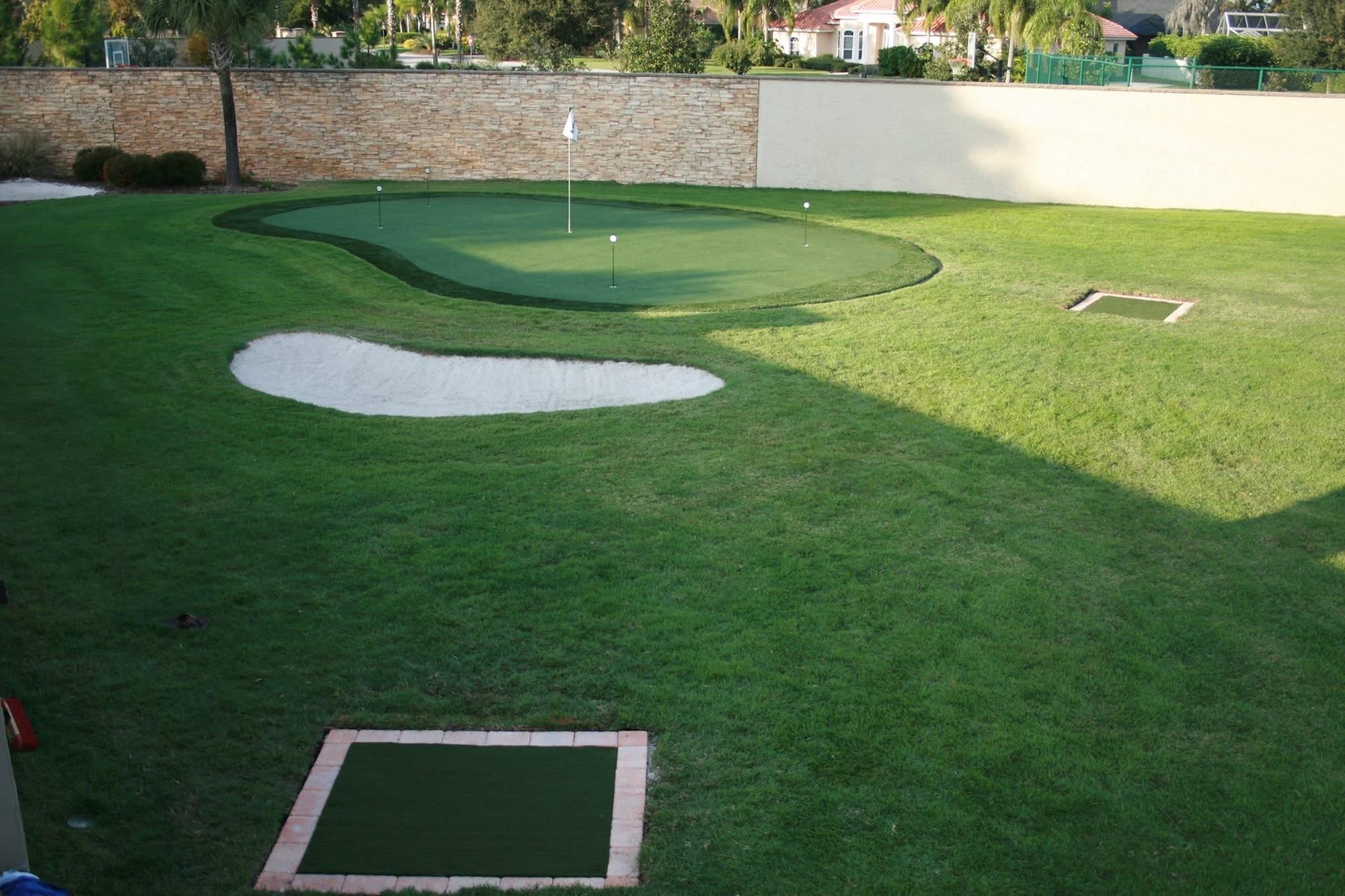 Putting green with a sand bunker and inset tee boxes inside a walled backyard
